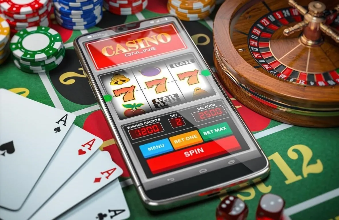 Phone with an online slot game on a casino table next to cards, chips and a roulette wheel
